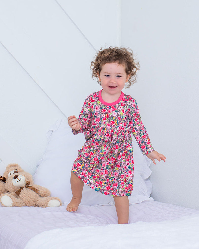Child in a floral dress standing on a bed with a teddy bear