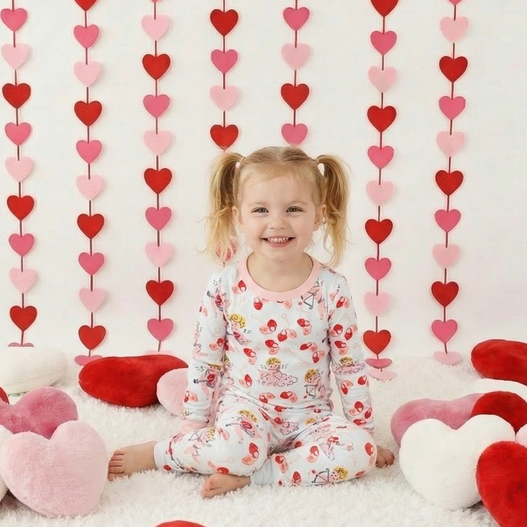 Child in heart-themed pajamas sitting on a white floor with heart decorations and a heart-patterned curtain.