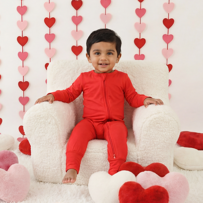 Child in a red outfit sitting on a white chair surrounded by heart-shaped decorations against a white wall.