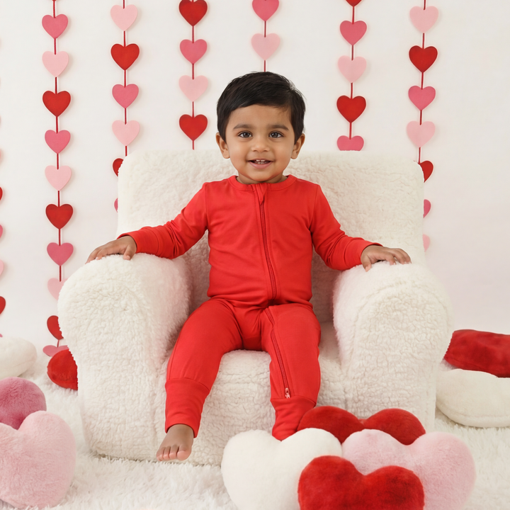 Child in a red outfit sitting on a white chair surrounded by heart-shaped decorations against a white wall.