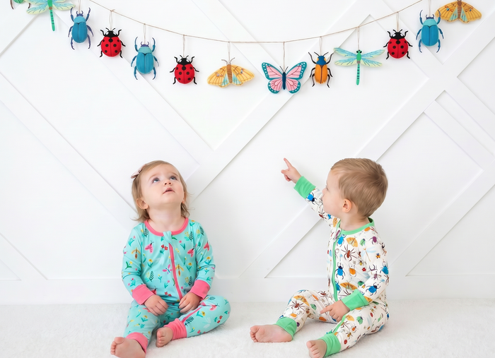 Two children in pajamas sitting on a white floor with a banner of colorful insect decorations above them.
