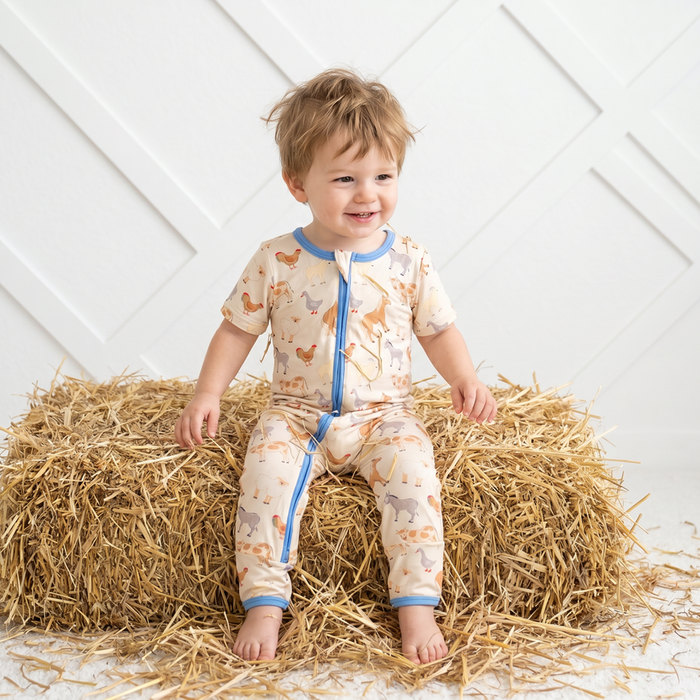Child wearing a patterned onesie sitting on a bale of hay against a white wall.