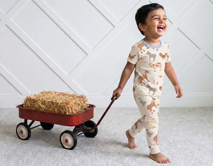 Child in pajamas with animal print pushing a red wagon filled with hay against a white paneled wall.