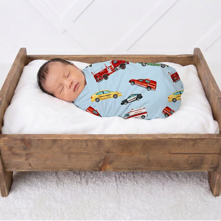 A sleeping newborn baby wrapped in a light blue swaddle blanket printed with colorful fire trucks and cars, resting in a wooden baby bed with white bedding against a white paneled wall.