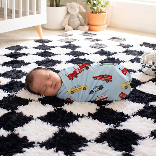 A baby sleeping on a black and white checkered shag rug in a bright nursery, wearing a light blue car-print swaddle. A wooden toy truck, a small disco ball, a crib, and plants are visible in the room.