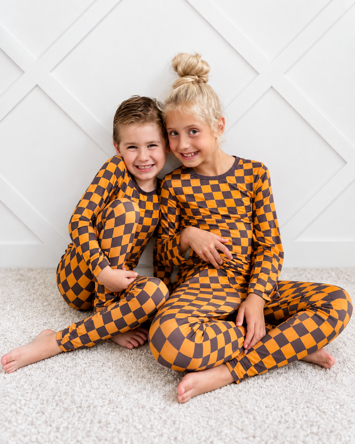 Two children sitting cross-legged on a light carpet, wearing matching long-sleeve pajamas with a mustard yellow and dark brown checkerboard pattern against a white paneled wall.