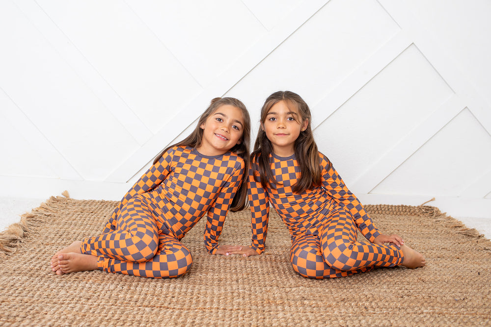 Two young girls sitting side-by-side on a woven jute rug, wearing matching orange and dark brown checkerboard patterned long-sleeve bodysuits against a white paneled wall.