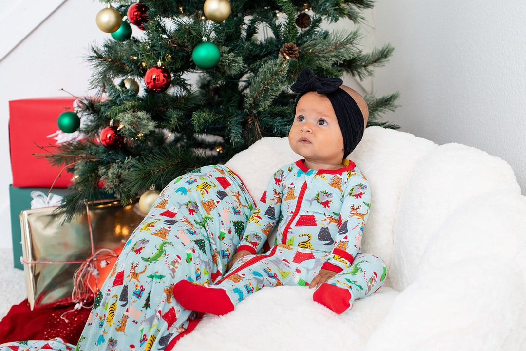 Baby in festive pajamas sitting on a white couch next to a decorated Christmas tree.