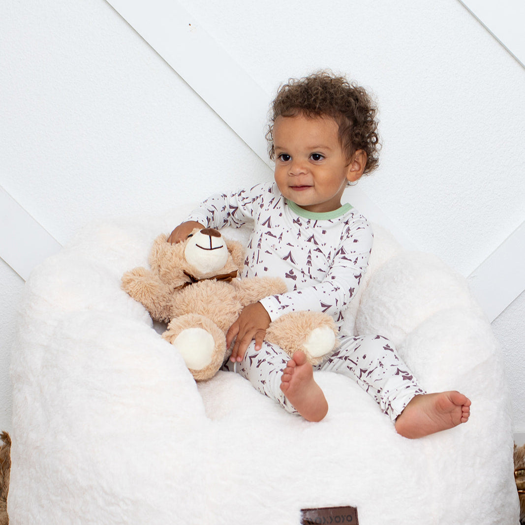 Child sitting on a white bean bag with a teddy bear, against a light background