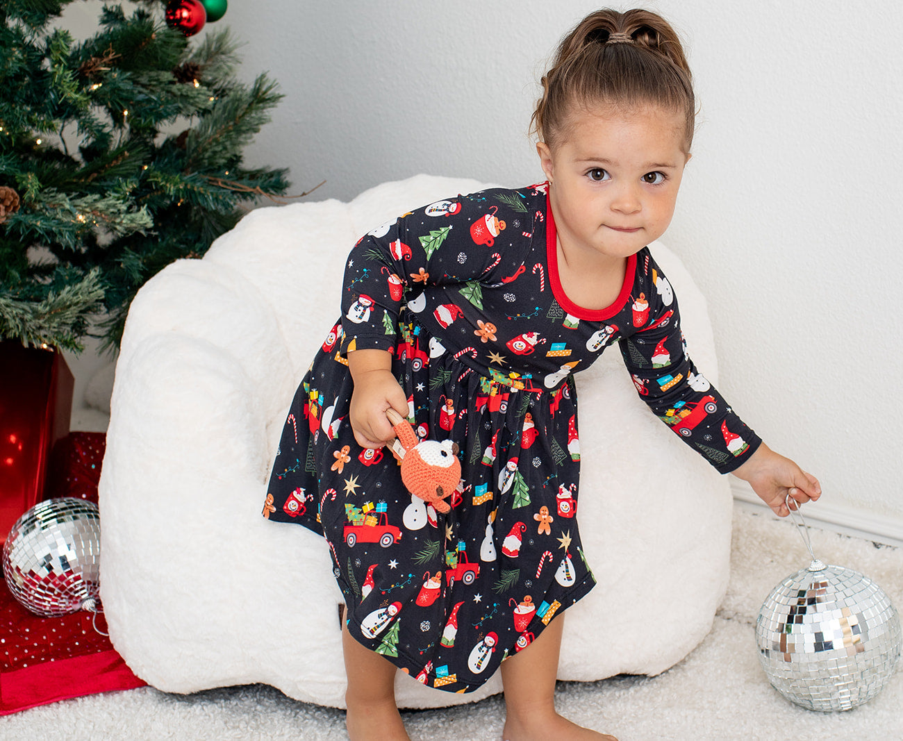Child in a festive dress sitting on a white chair with Christmas decorations in the background.