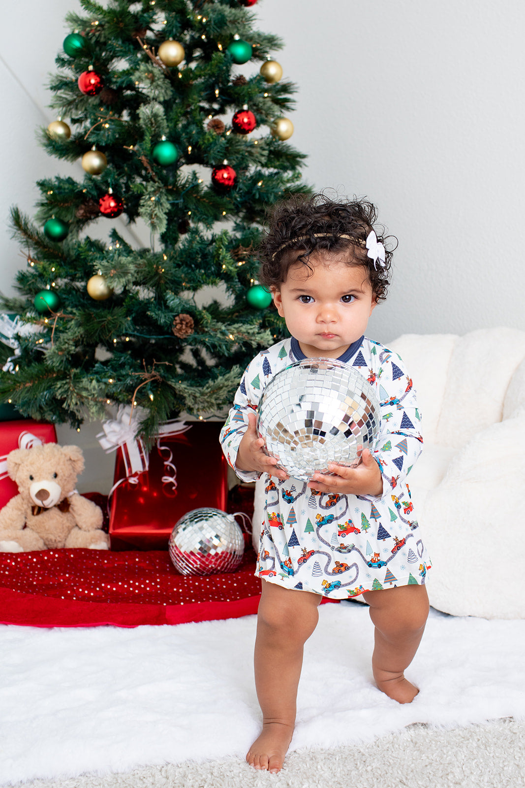 Child in pajamas standing in front of a decorated Christmas tree with a teddy bear and ornaments.