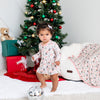 Baby in a floral dress standing in front of a decorated Christmas tree with presents and a white dog.