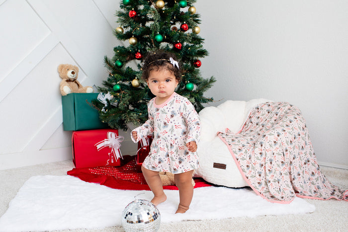 Baby in a floral dress standing in front of a decorated Christmas tree with presents and a white dog.