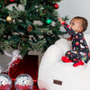 Baby in pajamas reaching for a Christmas tree with ornaments and presents underneath.
