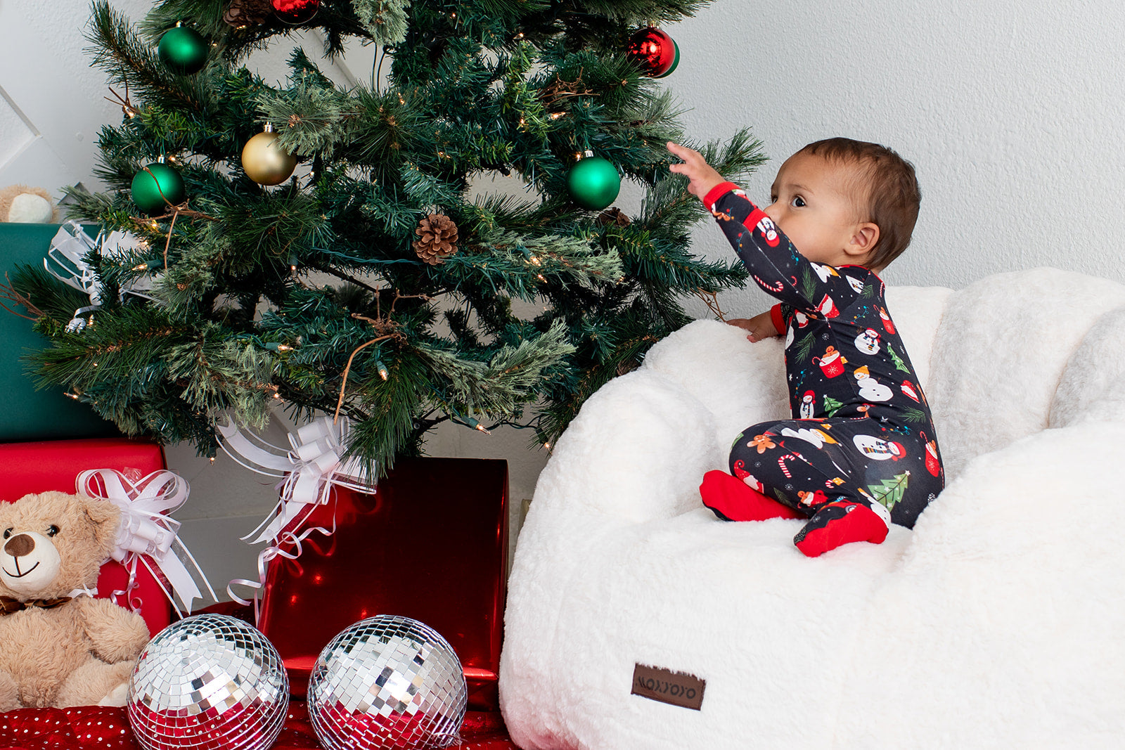Baby in pajamas reaching for a Christmas tree with ornaments and presents underneath.