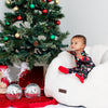 Child in pajamas sitting on a bean bag chair next to a decorated Christmas tree with presents and toys.