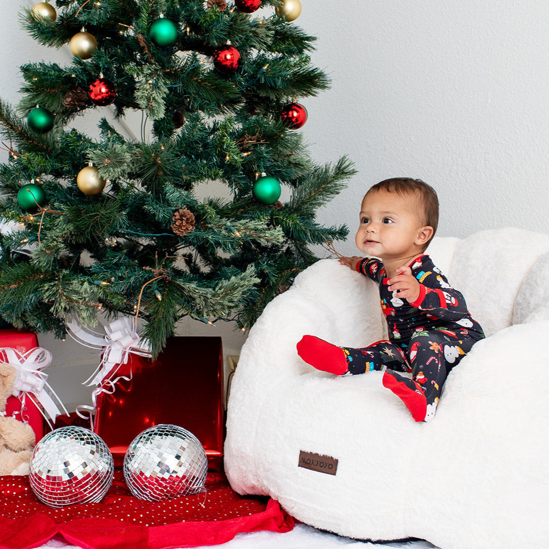 Child in pajamas sitting on a bean bag chair next to a decorated Christmas tree with presents and toys.
