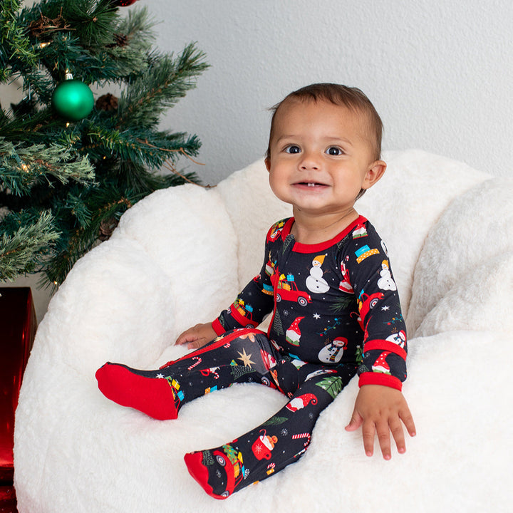 Baby in festive pajamas sitting on a bean bag chair next to a Christmas tree.