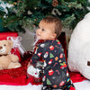 Child in festive pajamas sitting in front of a decorated Christmas tree with presents and a teddy bear.