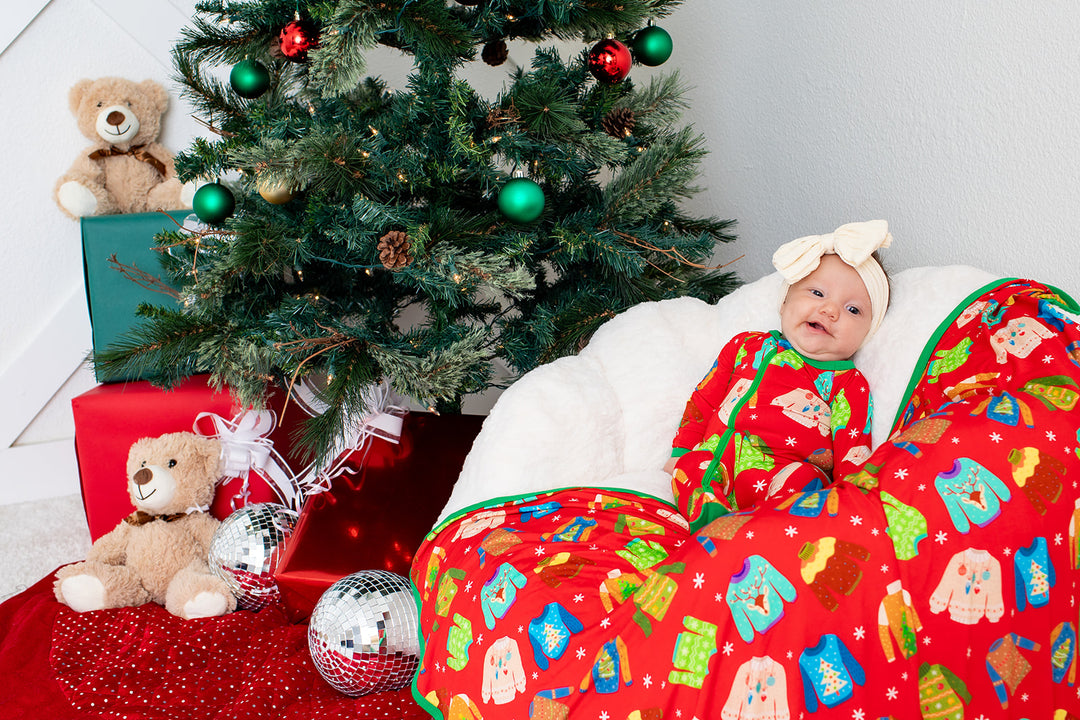 Baby in a colorful outfit sitting next to a Christmas tree with decorations and presents.