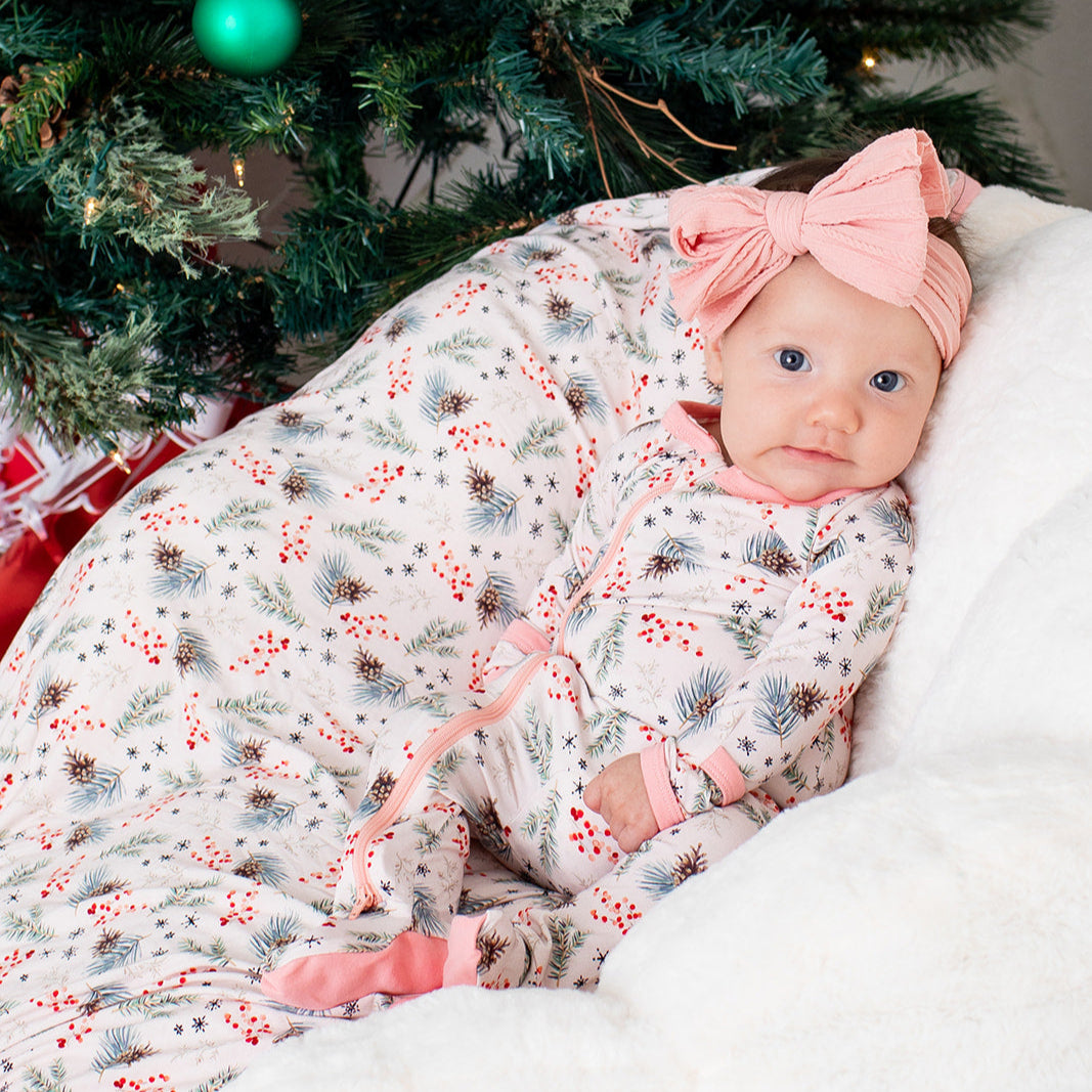 Baby wrapped in a floral blanket with a pink bow, sitting in front of a decorated Christmas tree.