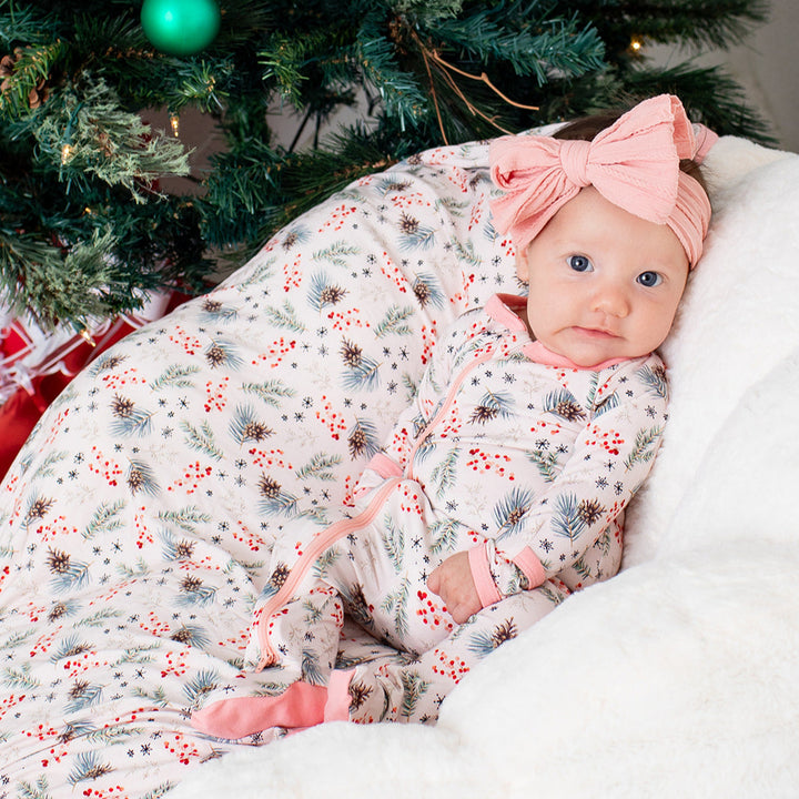 Baby wrapped in a floral blanket with a pink bow, sitting in front of a decorated Christmas tree.