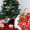Child in festive pajamas sitting next to a decorated Christmas tree with presents and teddy bears.