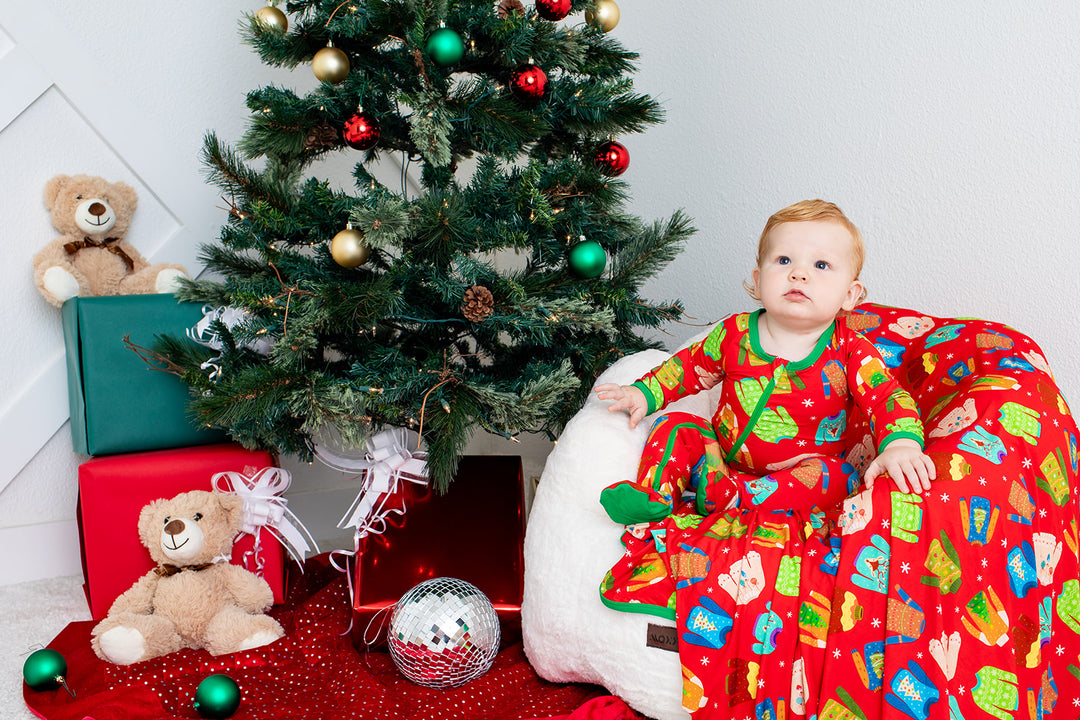 Child in festive pajamas sitting next to a decorated Christmas tree with presents and teddy bears.