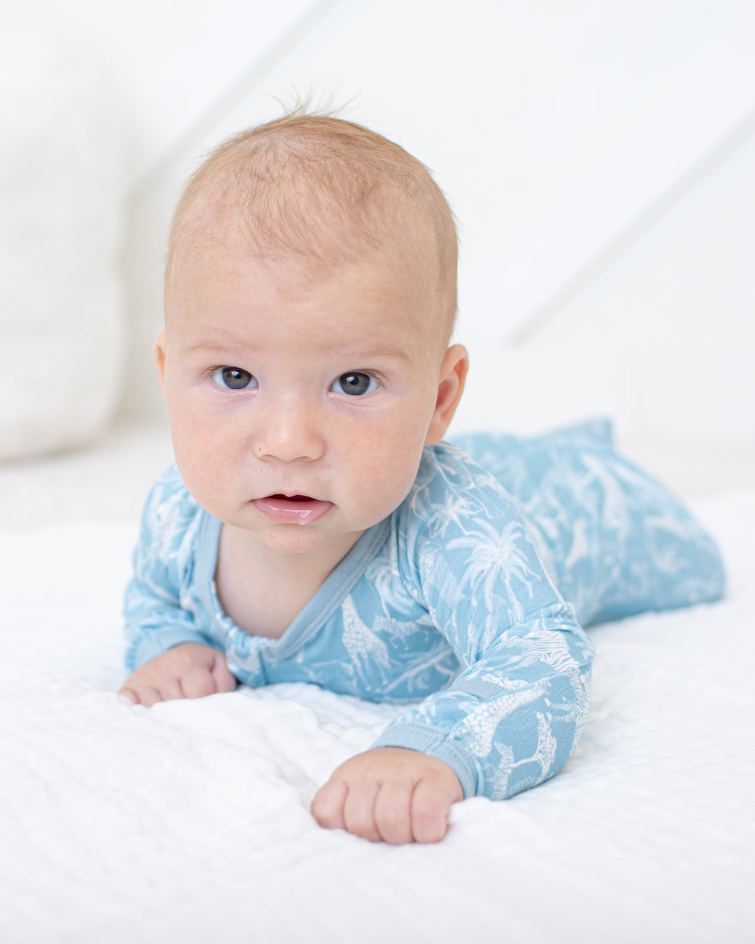 A baby lying on their stomach on a white textured blanket, wearing a light blue long-sleeve onesie with a white tropical leaf pattern.