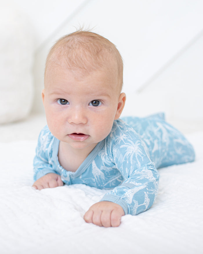 A baby lying on their stomach on a white textured blanket, wearing a light blue long-sleeve onesie with a white tropical leaf pattern.