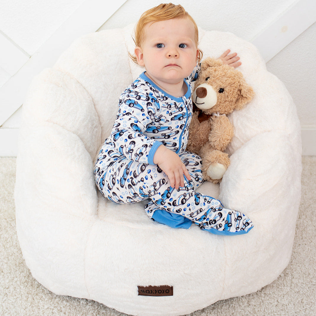 Baby sitting on a white bean bag chair holding a teddy bear in a room with white floor and wall.