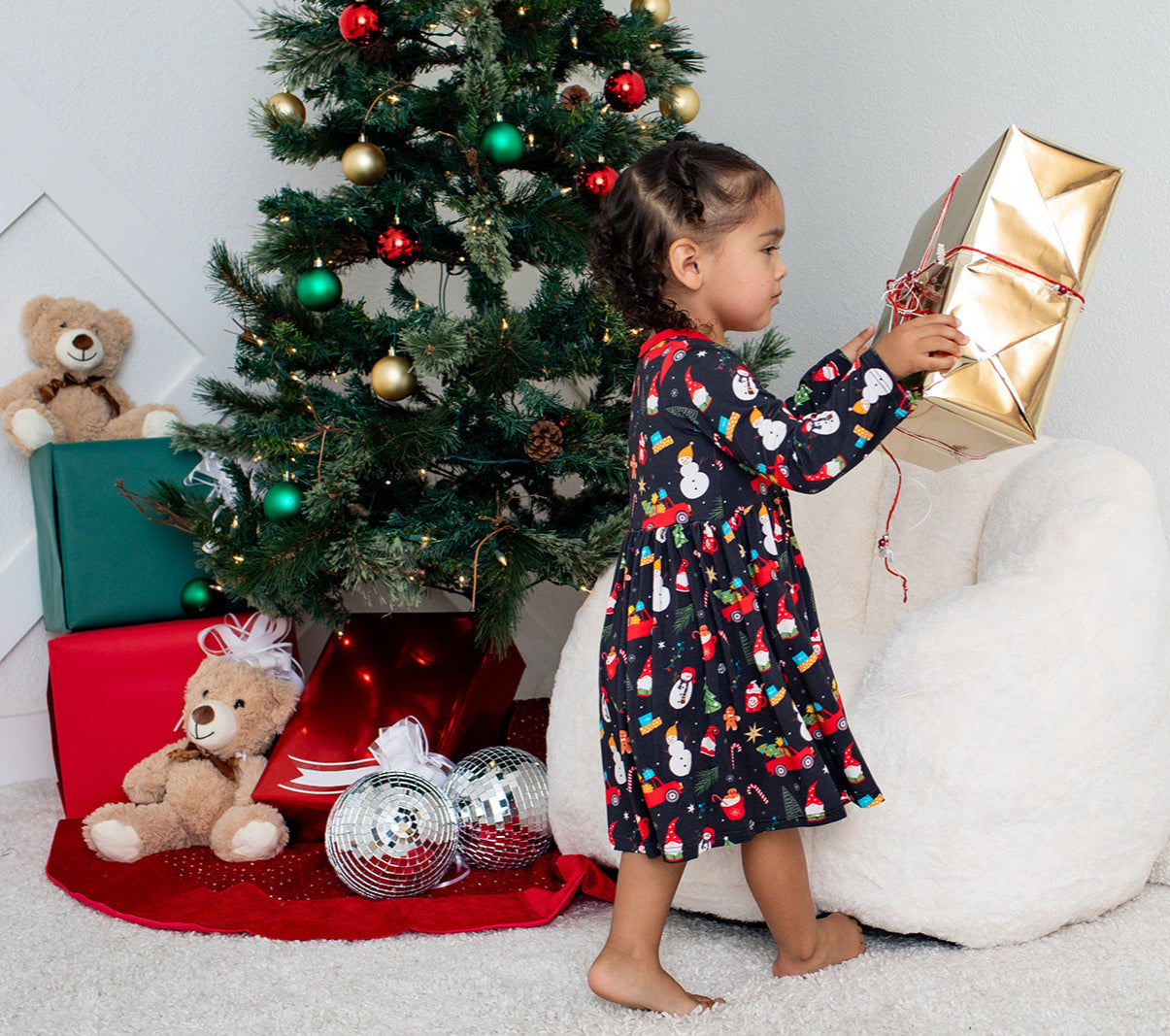 Child in a festive dress opening a gift near a decorated Christmas tree.