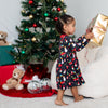 Child in a festive dress opening a gift near a decorated Christmas tree.