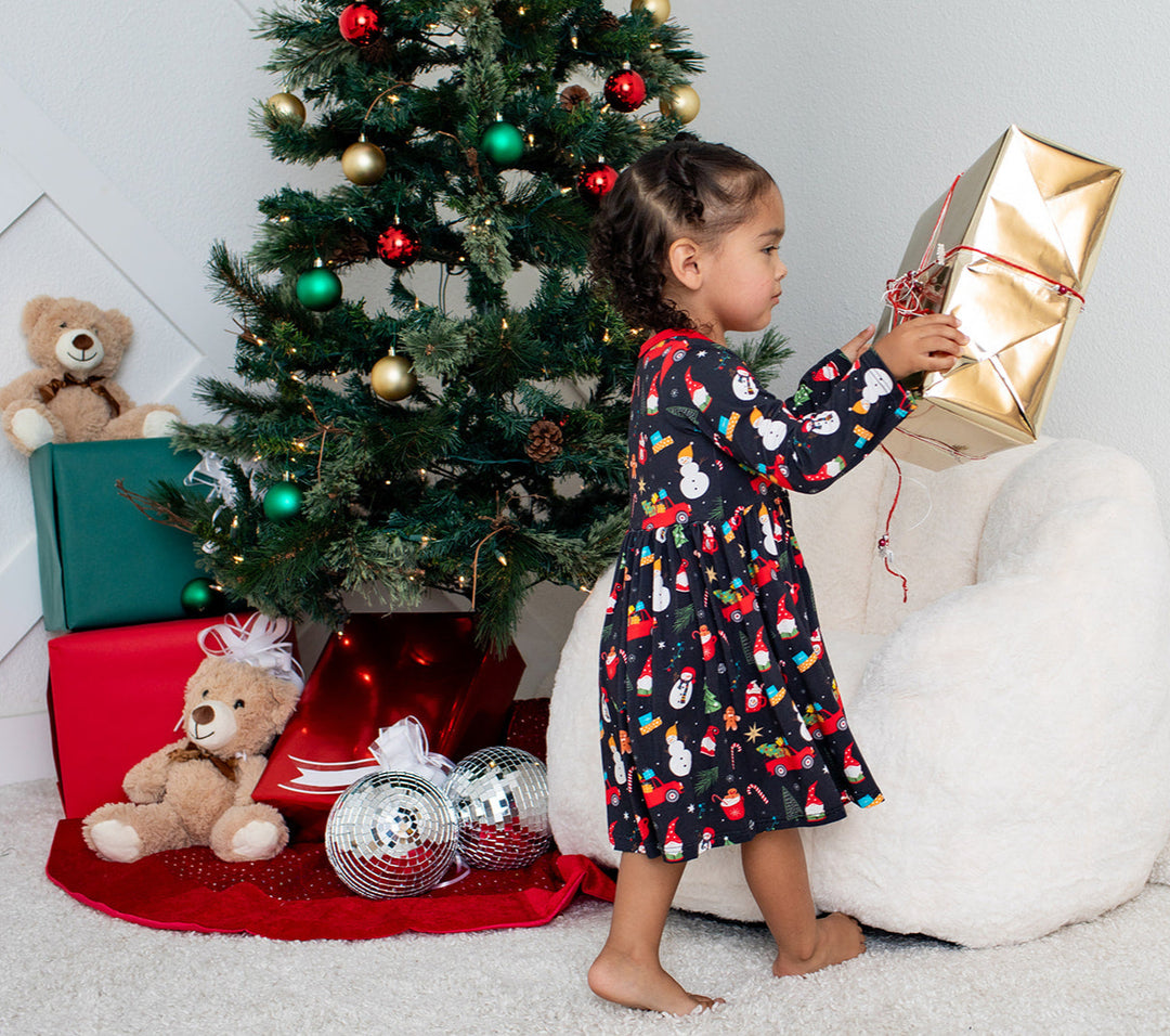 Child in a festive dress opening a gift near a decorated Christmas tree.