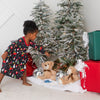 Child in pajamas standing next to a Christmas tree with presents and toys on a carpeted floor.