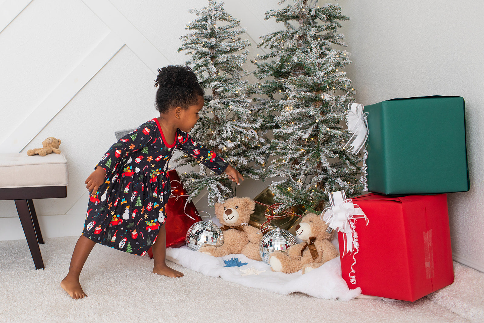 Child in pajamas standing next to a Christmas tree with presents and toys on a carpeted floor.