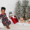 Child in a festive dress sitting on a white bean bag chair with Christmas decorations including a tree, presents, and teddy bears.