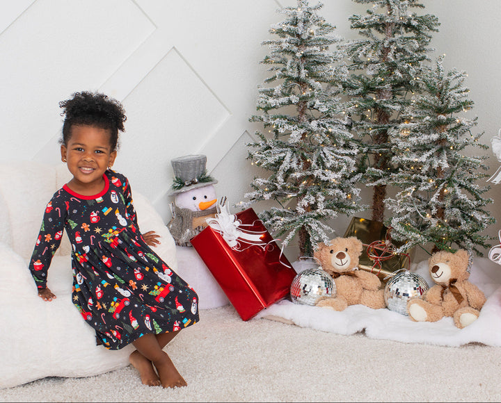 Child in a festive dress sitting on a white bean bag chair with Christmas decorations including a tree, presents, and teddy bears.