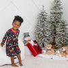 Child in pajamas standing next to Christmas trees and presents in a decorated room.