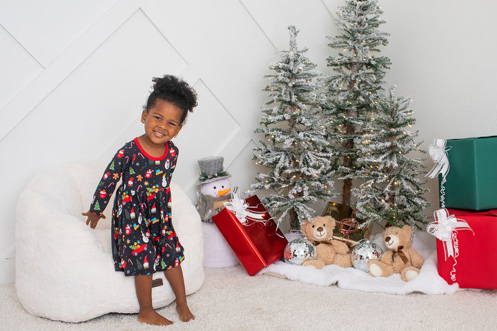 Child in pajamas standing next to Christmas trees and presents in a decorated room.