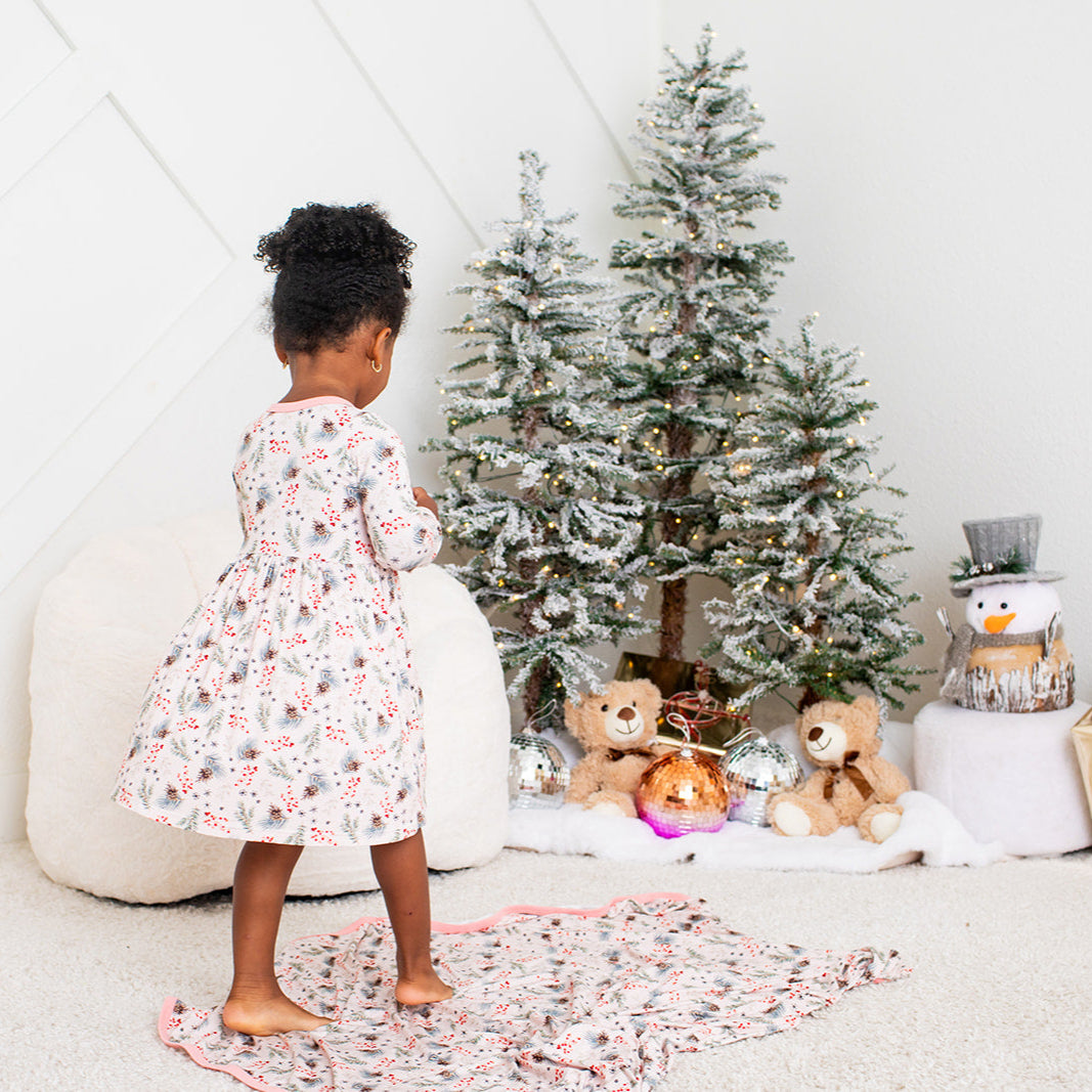 Child in a floral dress standing in front of a decorated Christmas tree with toys on a white floor.