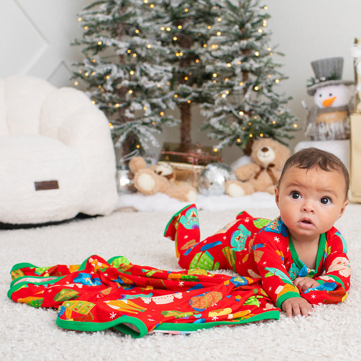 Baby in festive pajamas lying on a white carpet with Christmas decorations in the background.