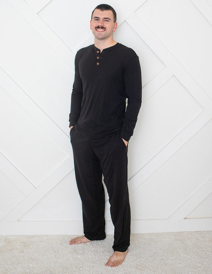 Man wearing a black long-sleeve shirt and pants standing against a white geometric-patterned wall.