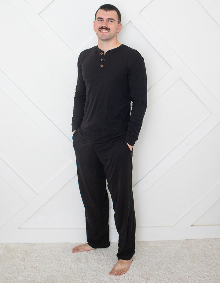 Man wearing a black long-sleeve shirt and pants standing against a white geometric-patterned wall.