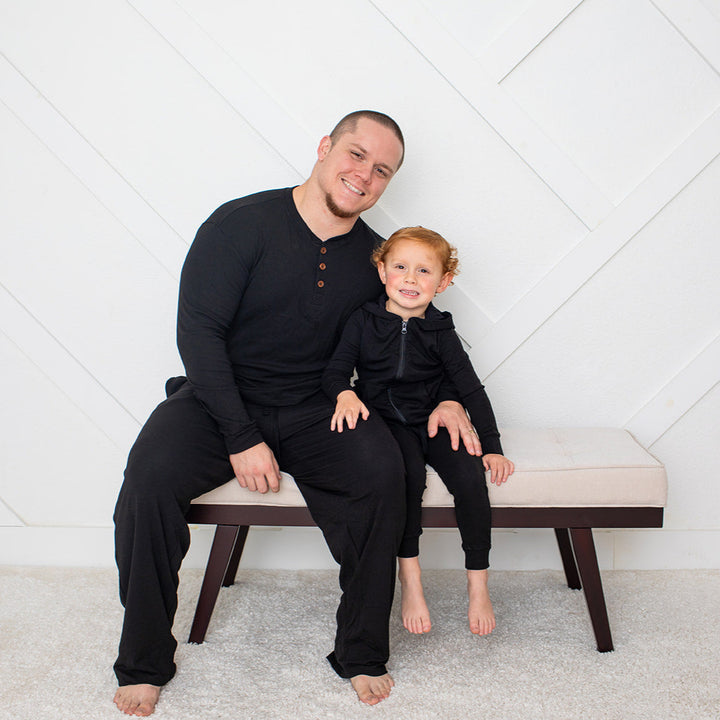 Man and child sitting on a bench wearing matching black outfits against a white geometric-patterned wall.