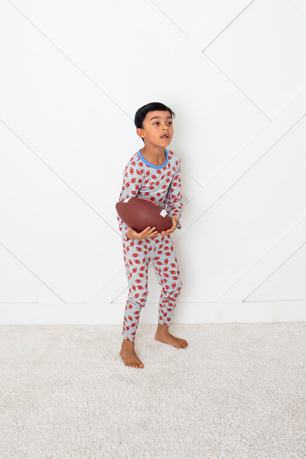 Child in red and white patterned pajamas holding a brown football on a white floor.