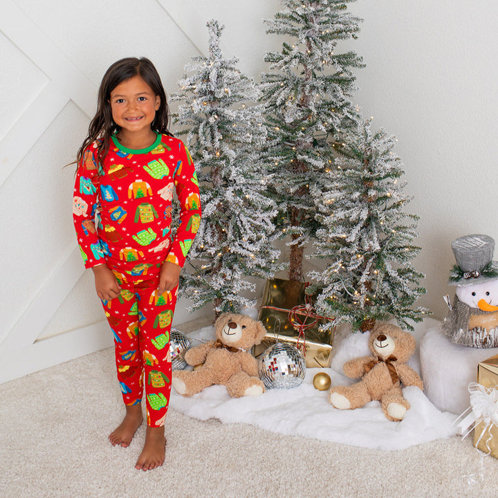 Child wearing red Christmas pajamas standing in front of a decorated Christmas tree with teddy bears and presents.