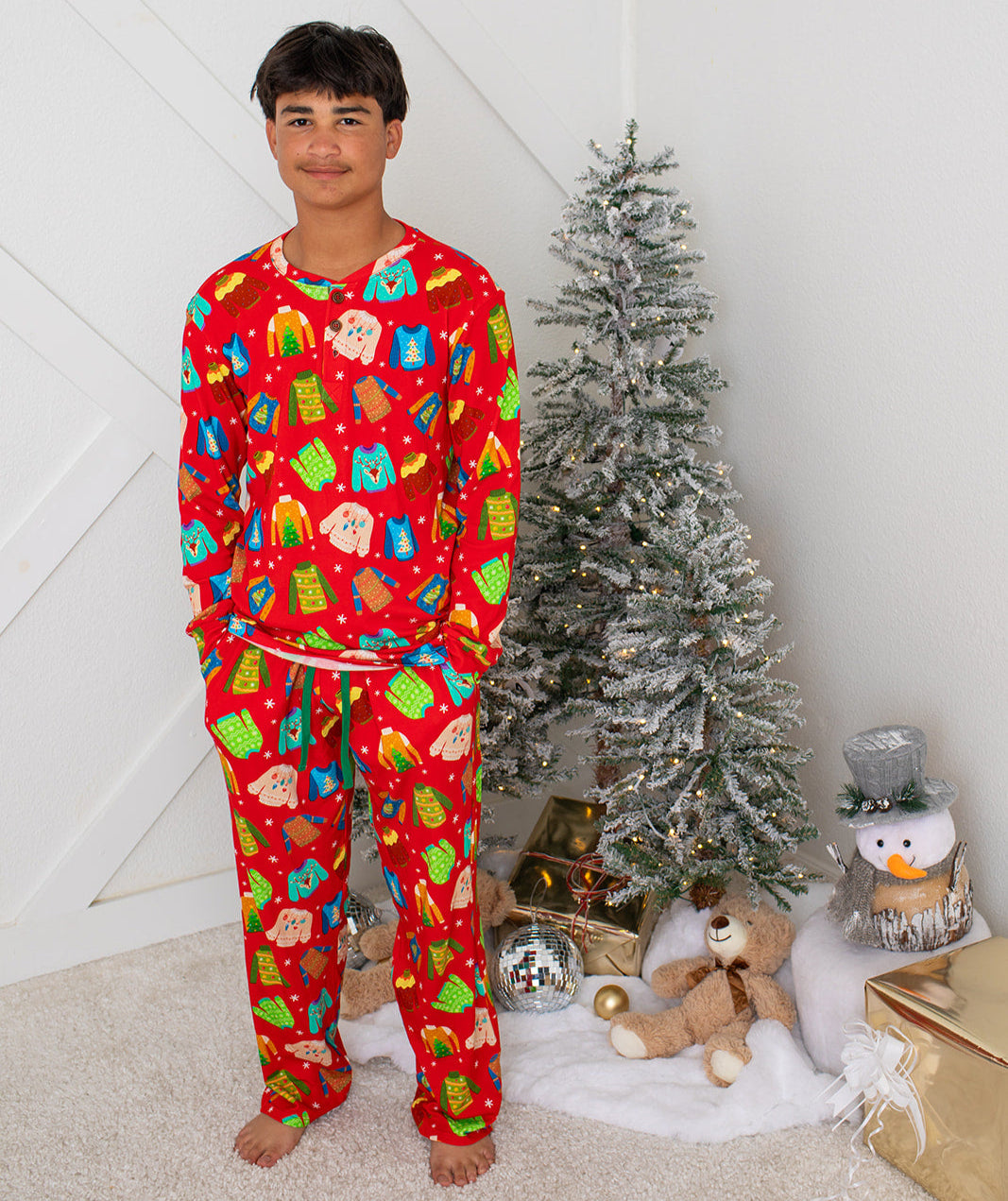 Person wearing a red Christmas-themed pajama set standing next to a decorated tree with ornaments and a teddy bear.