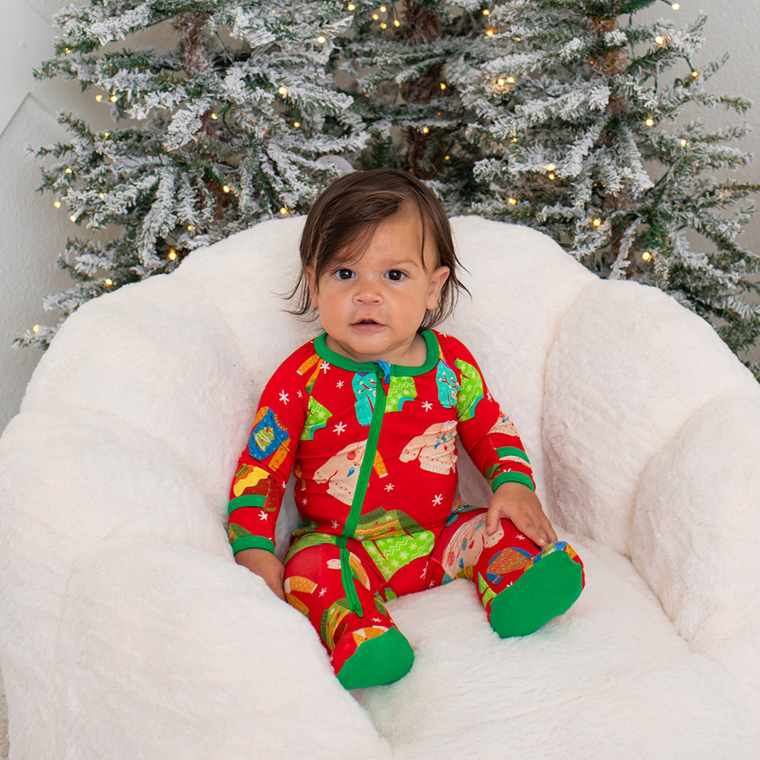 Baby in festive pajamas sitting on a white bean bag chair with a decorated Christmas tree in the background.