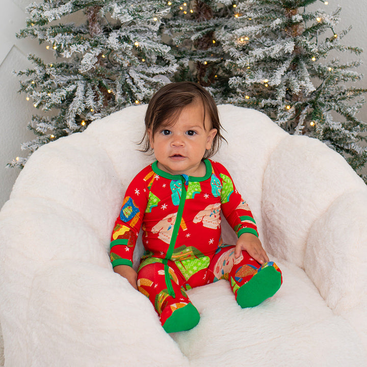Baby in festive pajamas sitting on a white bean bag chair with a decorated Christmas tree in the background.